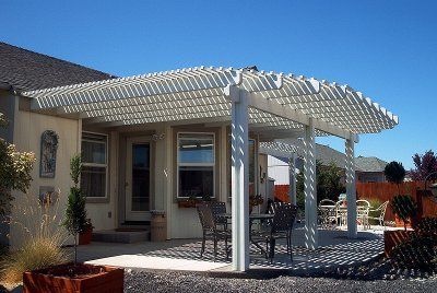 White pergola over a patio with outdoor furniture, attached to a light-colored house under a clear blue sky.