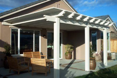 White pergola covers a patio with wood furniture; adjacent house is light brown.