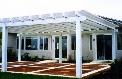 White pergola over a patio with a house in the background. Brown and white patio. Green grass.