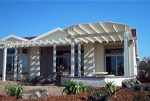 Beige house with pergola and large windows, surrounded by landscaping and blue sky.