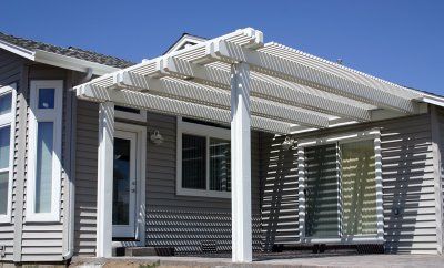 White pergola attached to a gray house, casting shadows. Includes glass door and sliding door. Sunny day.