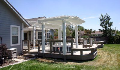 Wooden deck with white pergola, attached to a light blue house, surrounded by green grass under a clear blue sky.
