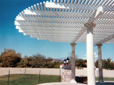 White pergola with slatted roof and column supports, set outdoors with blue sky and trees.