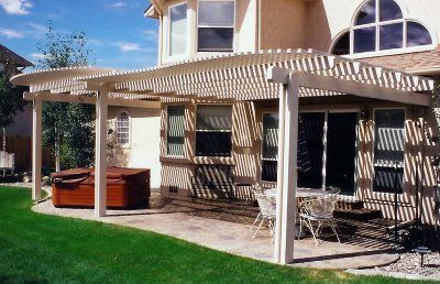 Beige pergola over a patio with a hot tub and table, attached to a house with a curved window.