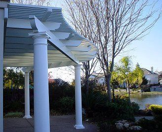 White pergola with columns by a lake, with trees and a house in the background.