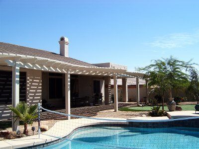 Patio with pergola adjacent to a house, pool in foreground, blue sky.