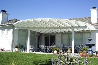 White pergola covering a patio with outdoor furniture, beside a white house, blue sky.