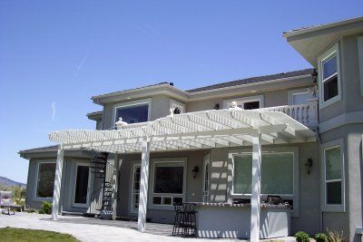 A white pergola shades a back patio of a two-story gray house under a blue sky.