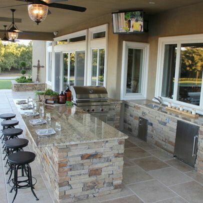Outdoor kitchen with bar seating, grill, and TV. Stone and granite construction, overlooking a backyard.