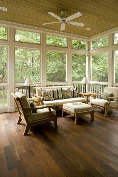 Sunroom with wooden floors, light-colored furniture, large windows, and a ceiling fan. Overlooks a lush green outdoor view.