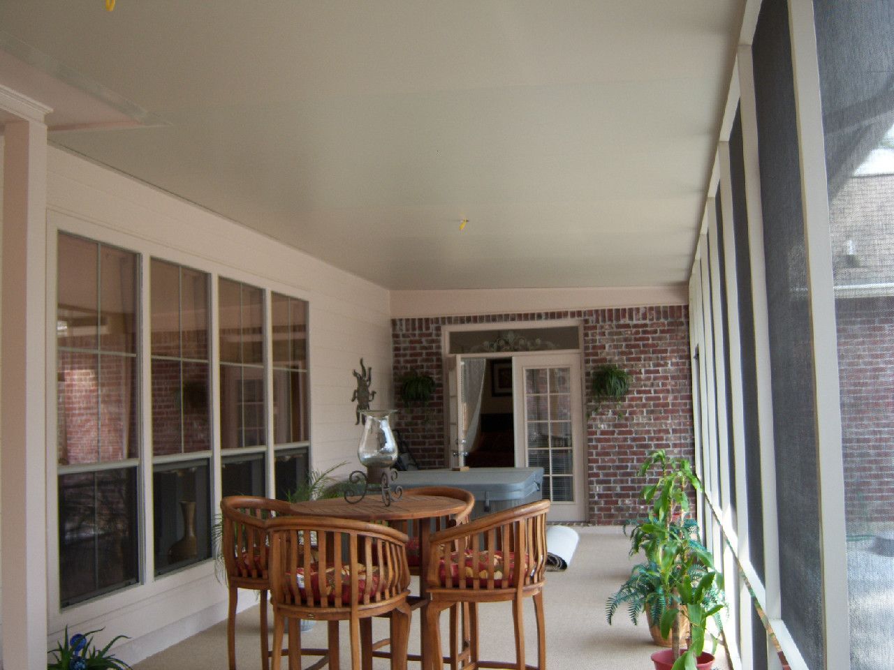 Screened porch with a table, chairs, and plants; windows on one side, brick wall with a door on the other.