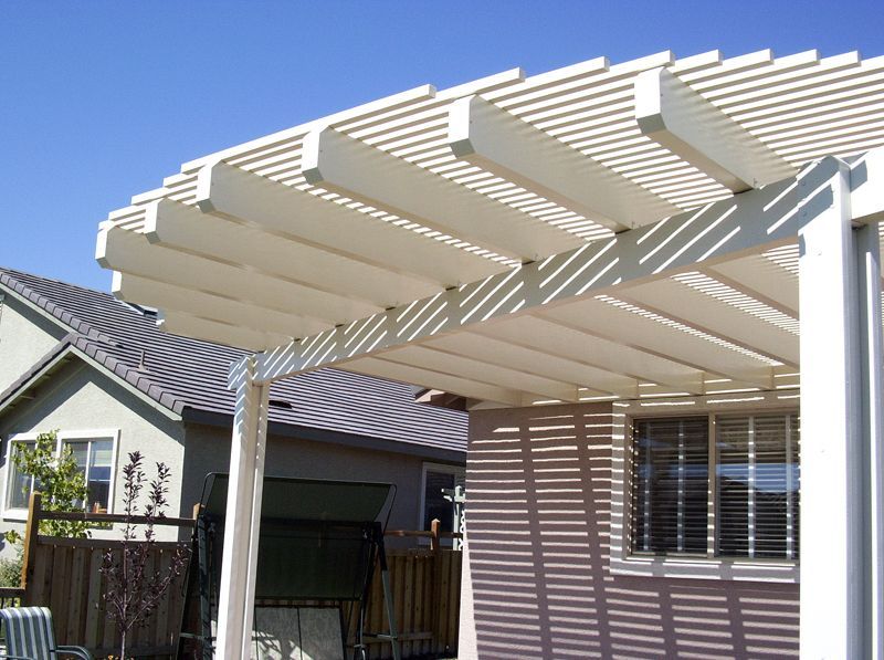 White pergola casting shadows on a beige house with a clear blue sky in the background.