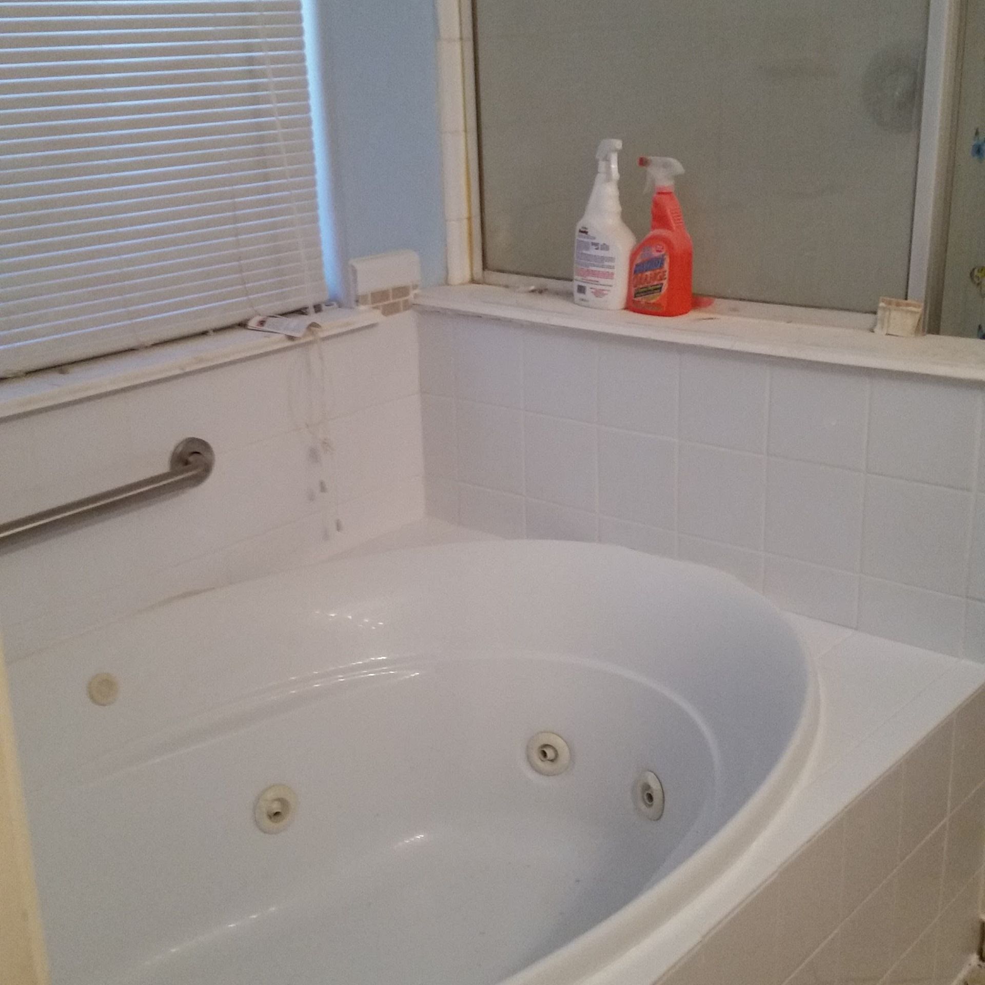 White jacuzzi tub with a grab bar, next to tiled wall with a window, and cleaning supplies