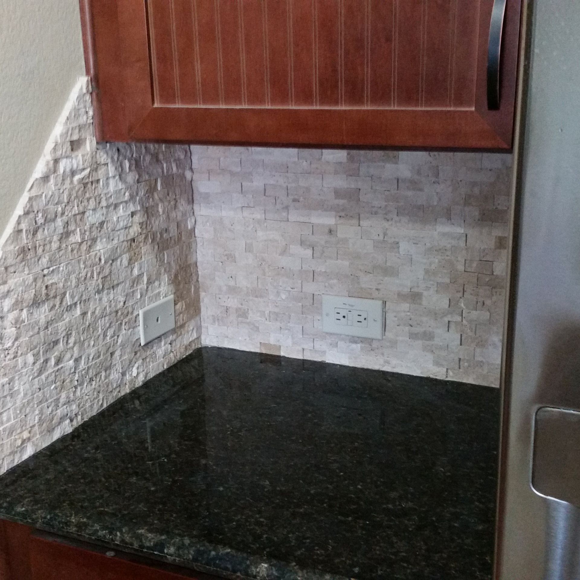 Kitchen corner with stone backsplash, dark granite countertop, wooden cabinets, and electrical outlet