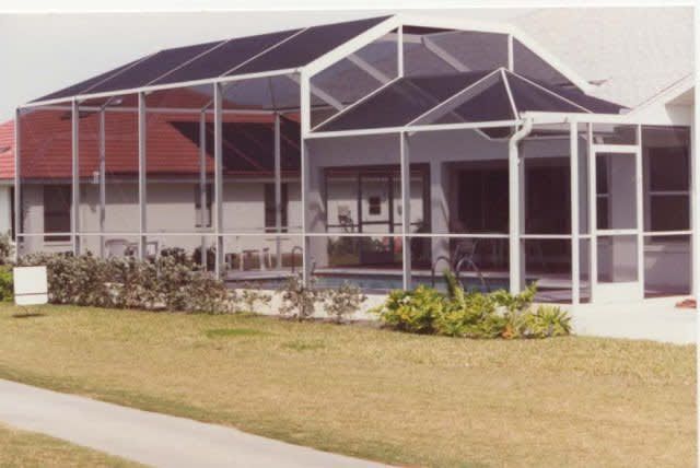 Screened-in pool enclosure with white framing, attached to a house with red roof tiles and a green lawn