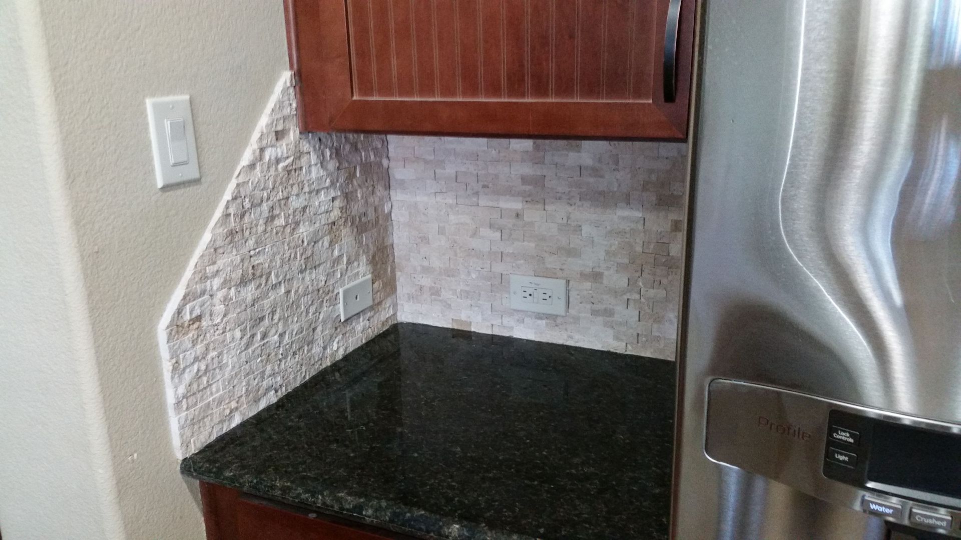 Kitchen counter with granite top, tiled backsplash, wooden cabinets, and stainless steel refrigerator
