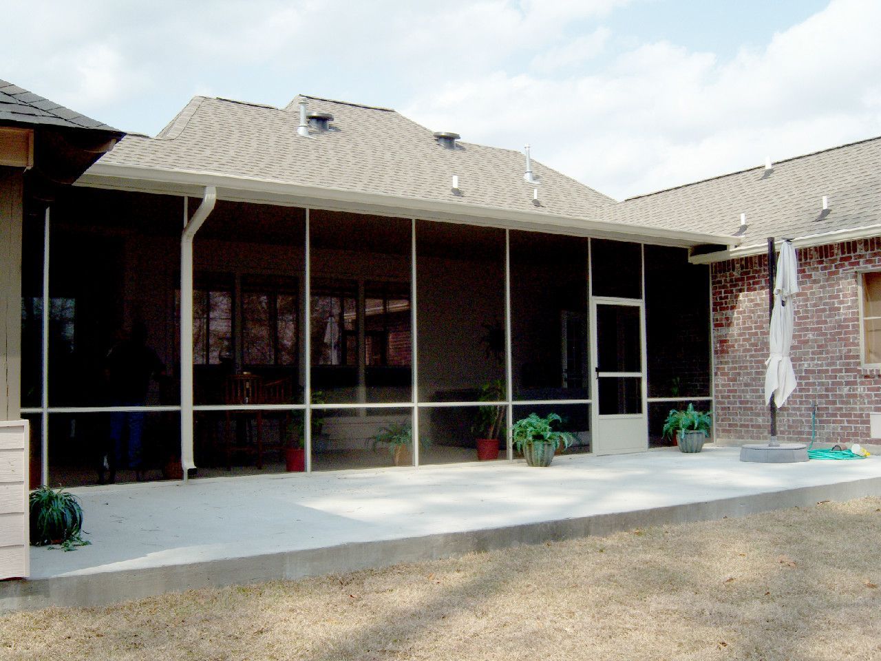 Screened-in patio with concrete flooring, attached to a brick house, overlooking a yard, and under a brown roof.
