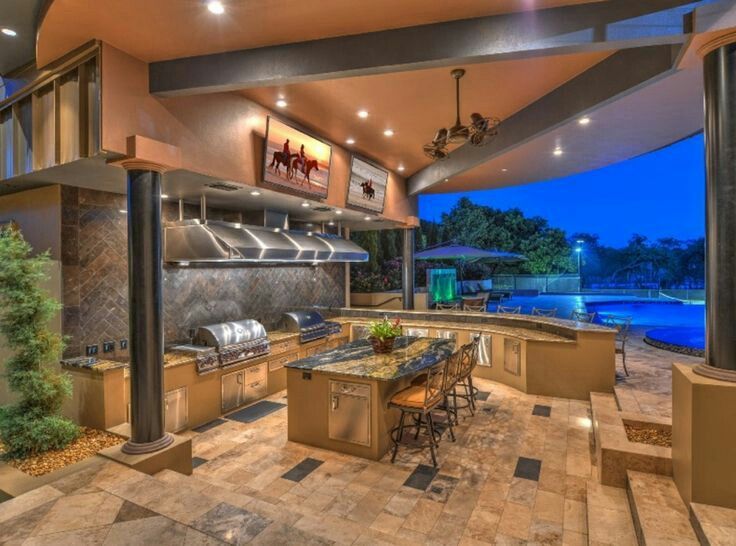 Outdoor kitchen with grills, bar, and seating area, overlooking a pool at dusk.