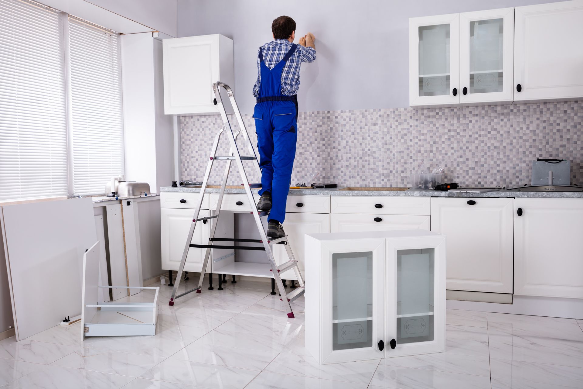 Person on a ladder installing a cabinet in a white kitchen, with components and tools nearby