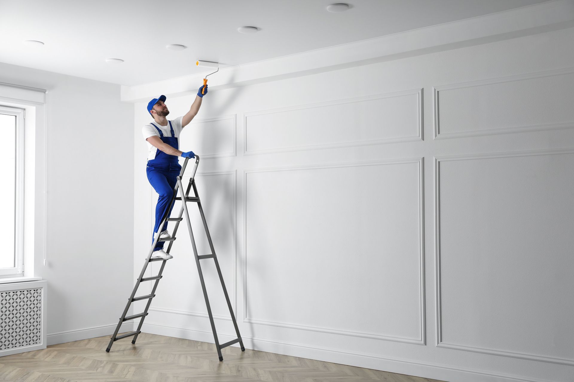 A person in blue overalls paints a white ceiling trim from a ladder in a white room