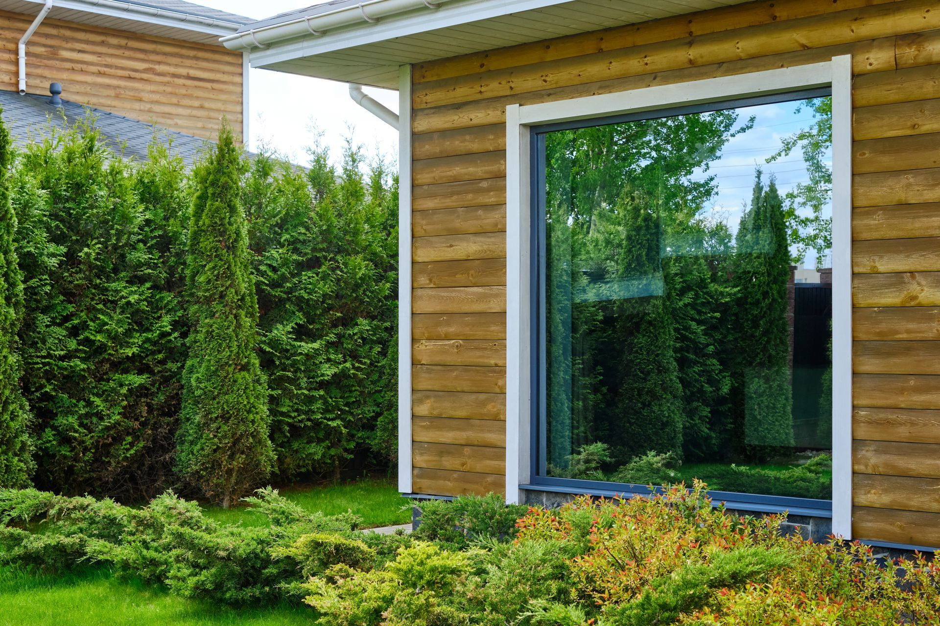 Exterior of a building with a large window reflecting green trees and landscaping, surrounded by wooden siding.