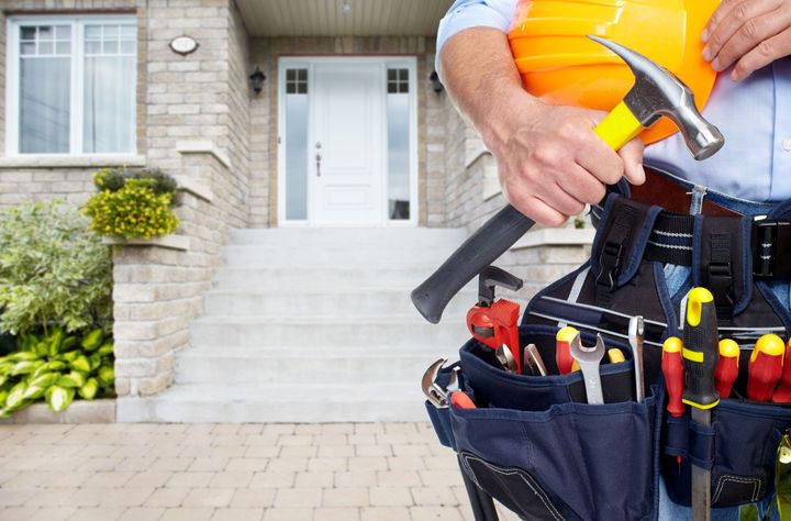 Handyman with tool belt holding hammer and hard hat in front of a house