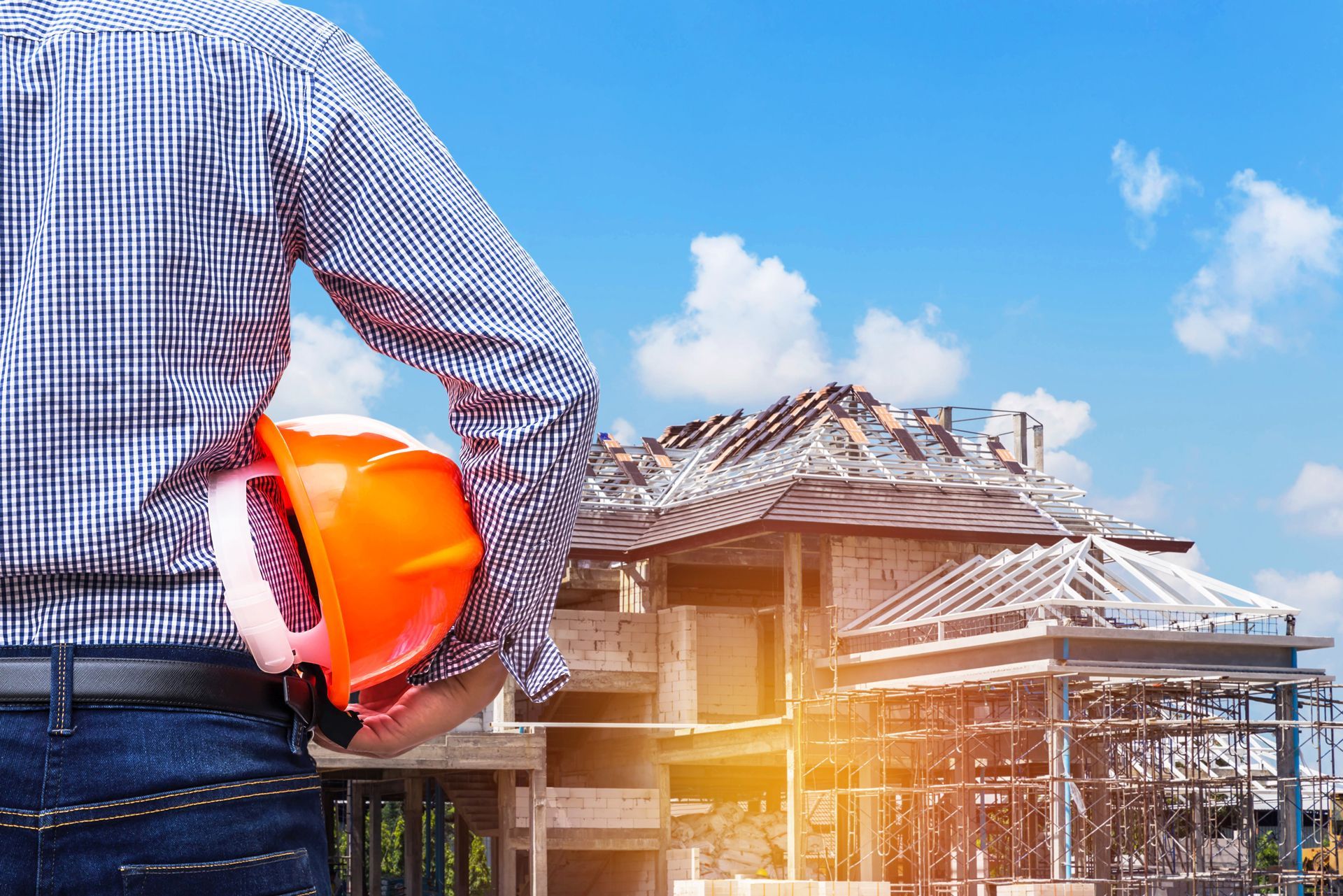 Construction worker overlooking a building under construction, holding orange hard hat; blue sky in background