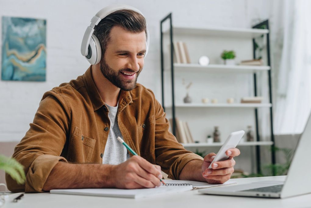Una persona con auriculares sonríe mientras escribe en un cuaderno y sostiene un teléfono inteligente 