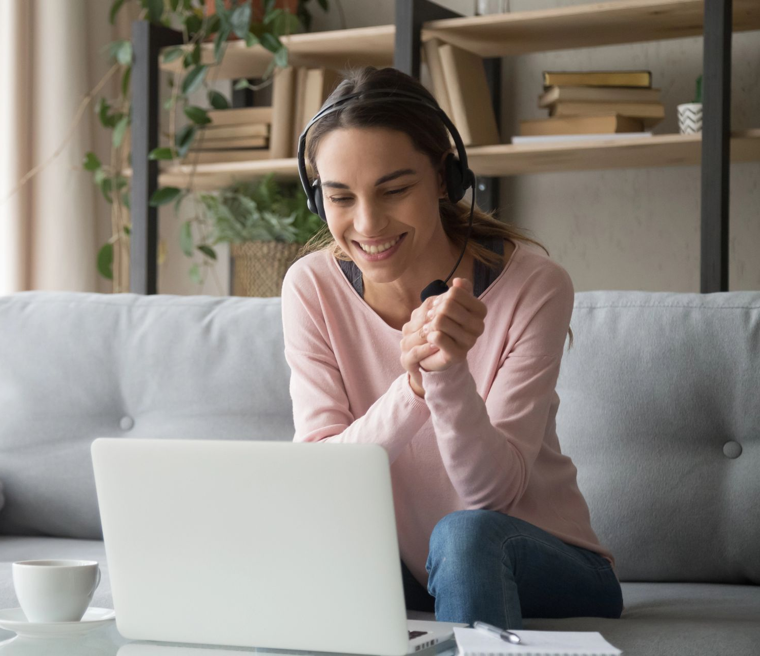 Una persona sonriente, con auriculares puestos, está sentada en un sofá gris, mirando una computadora
