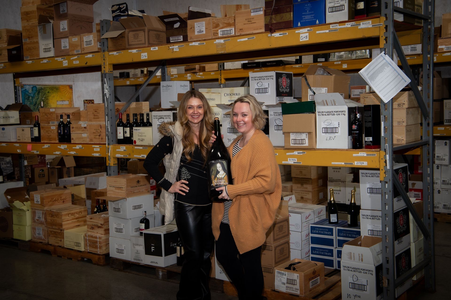 two women are standing in a warehouse holding bottles of wine .