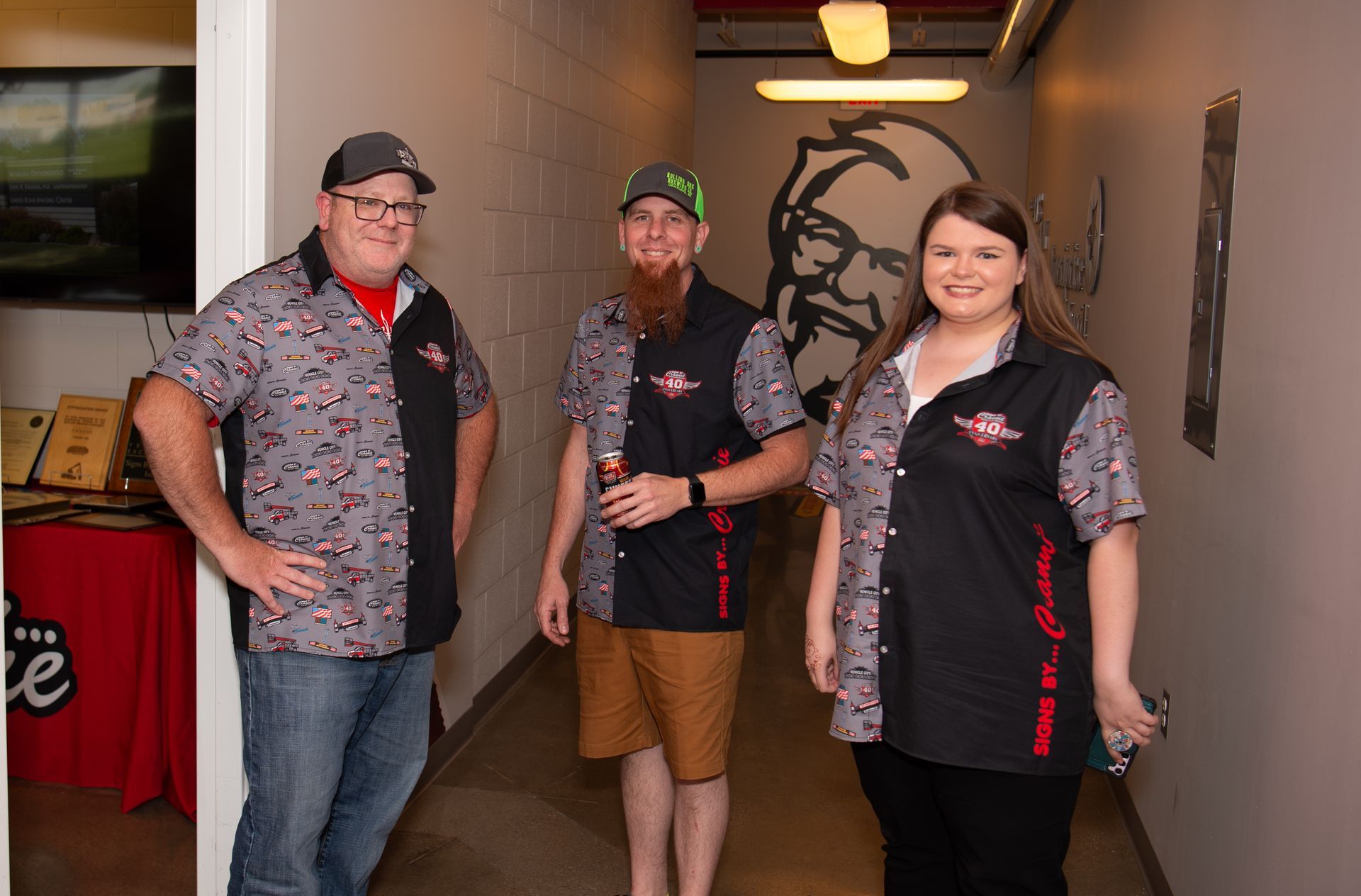 Three people are standing in a hallway with a kfc logo on the wall.
