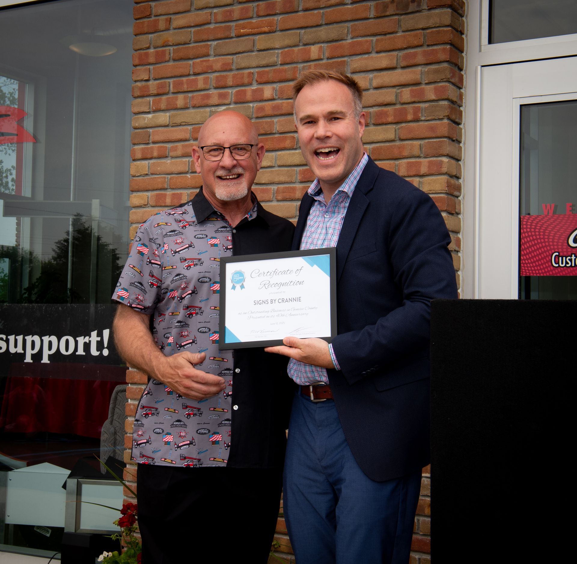 Two men holding a certificate in front of a building that says support