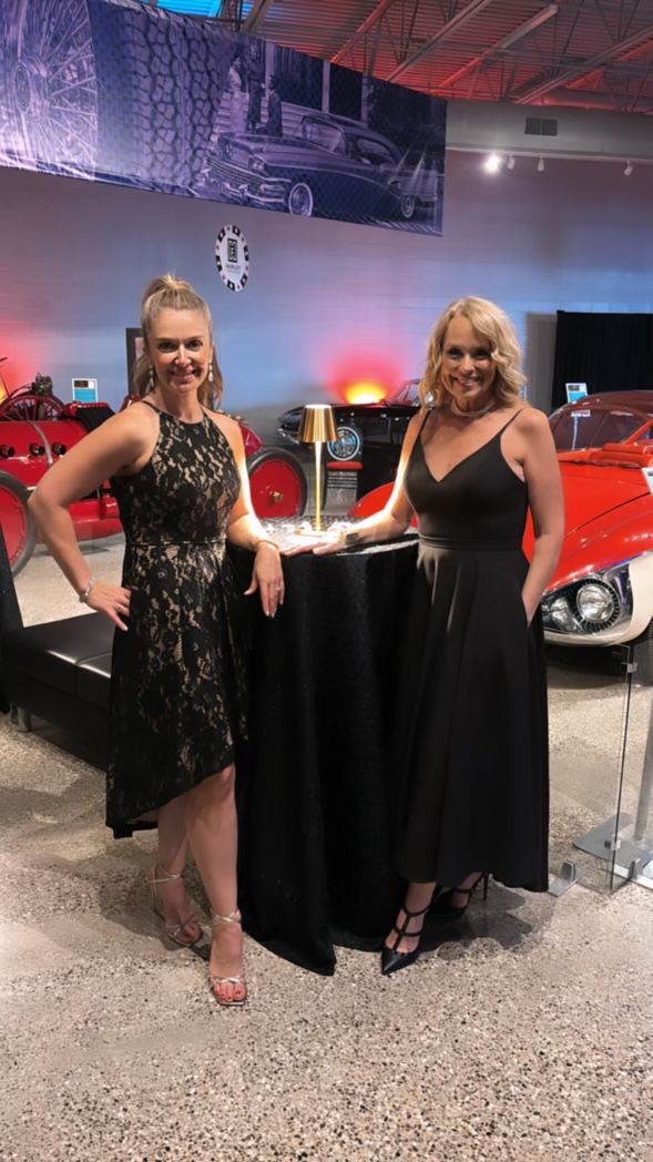 Two women in black dresses are standing next to each other in front of a red car.