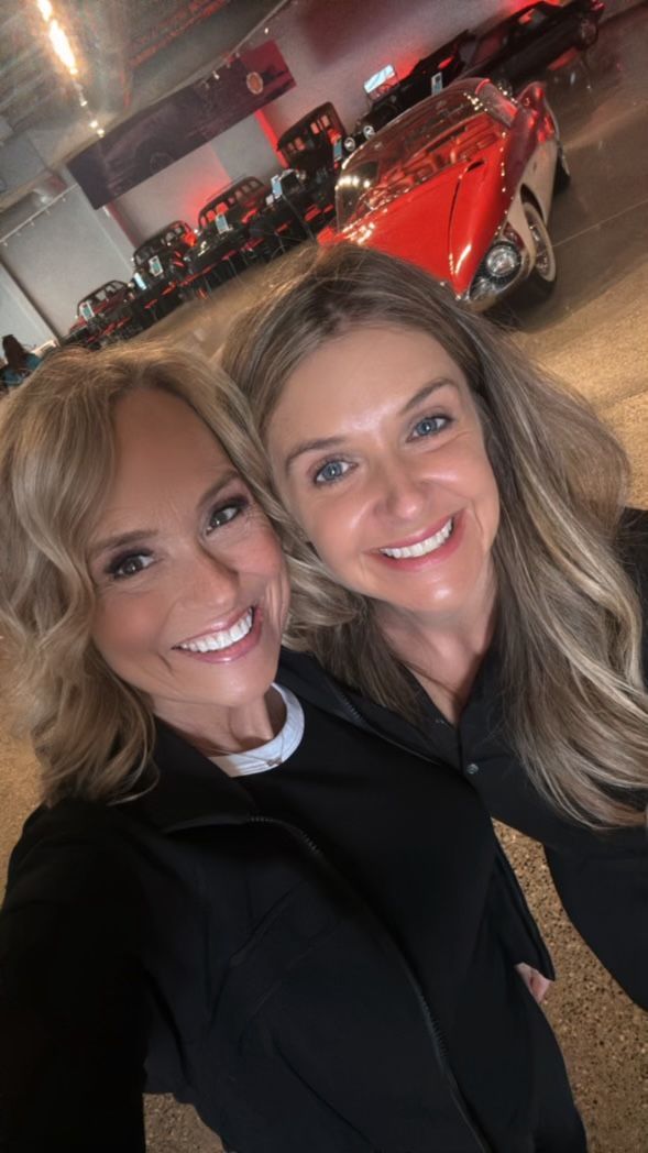 Two women are posing for a picture in front of a red car.