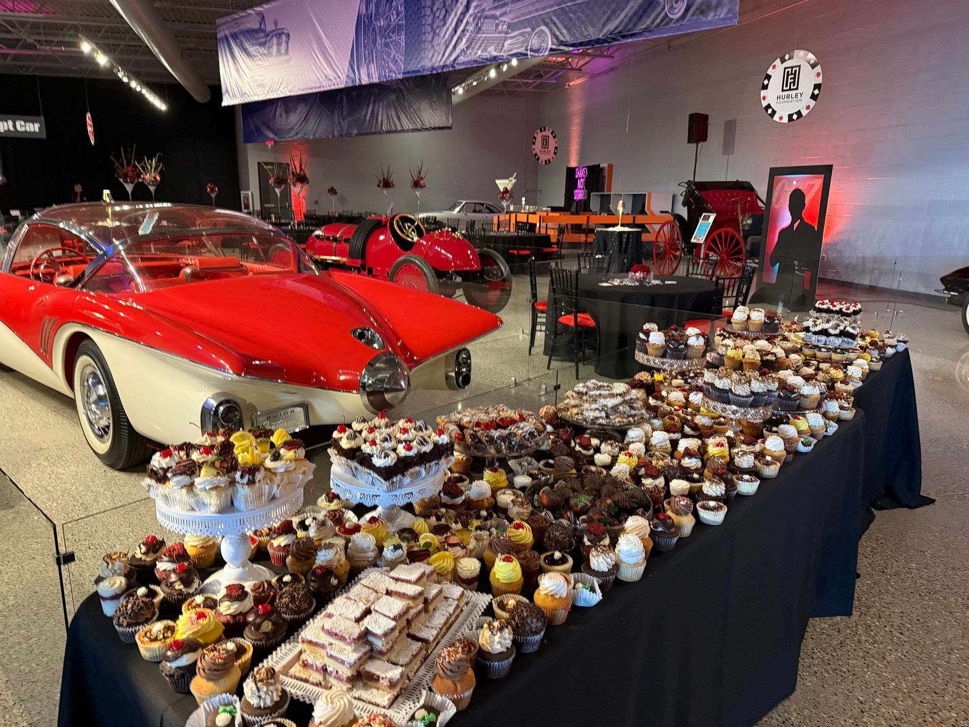 A table filled with cupcakes and a car in the background.