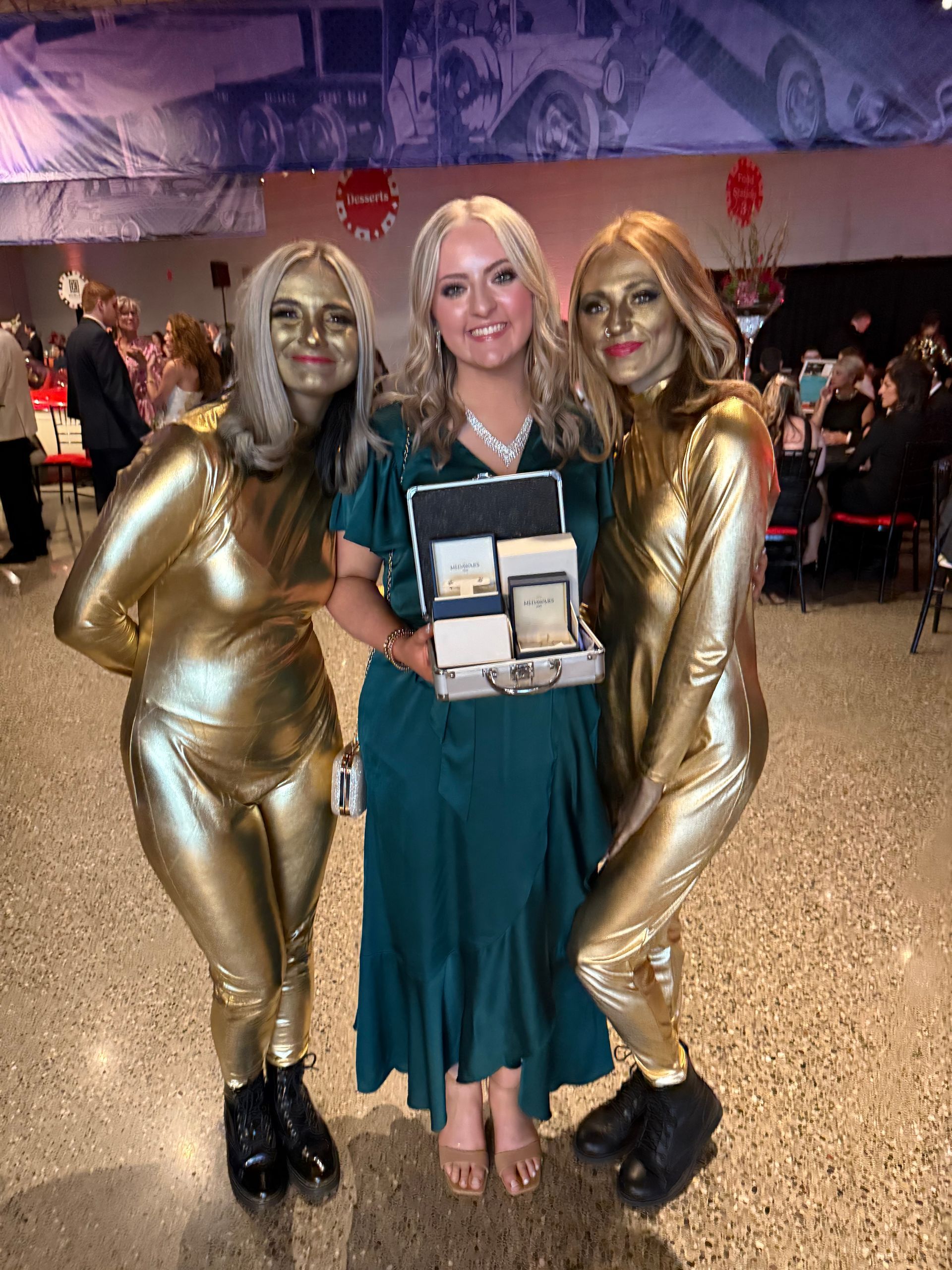 A woman in a green dress is standing next to two women in gold costumes.