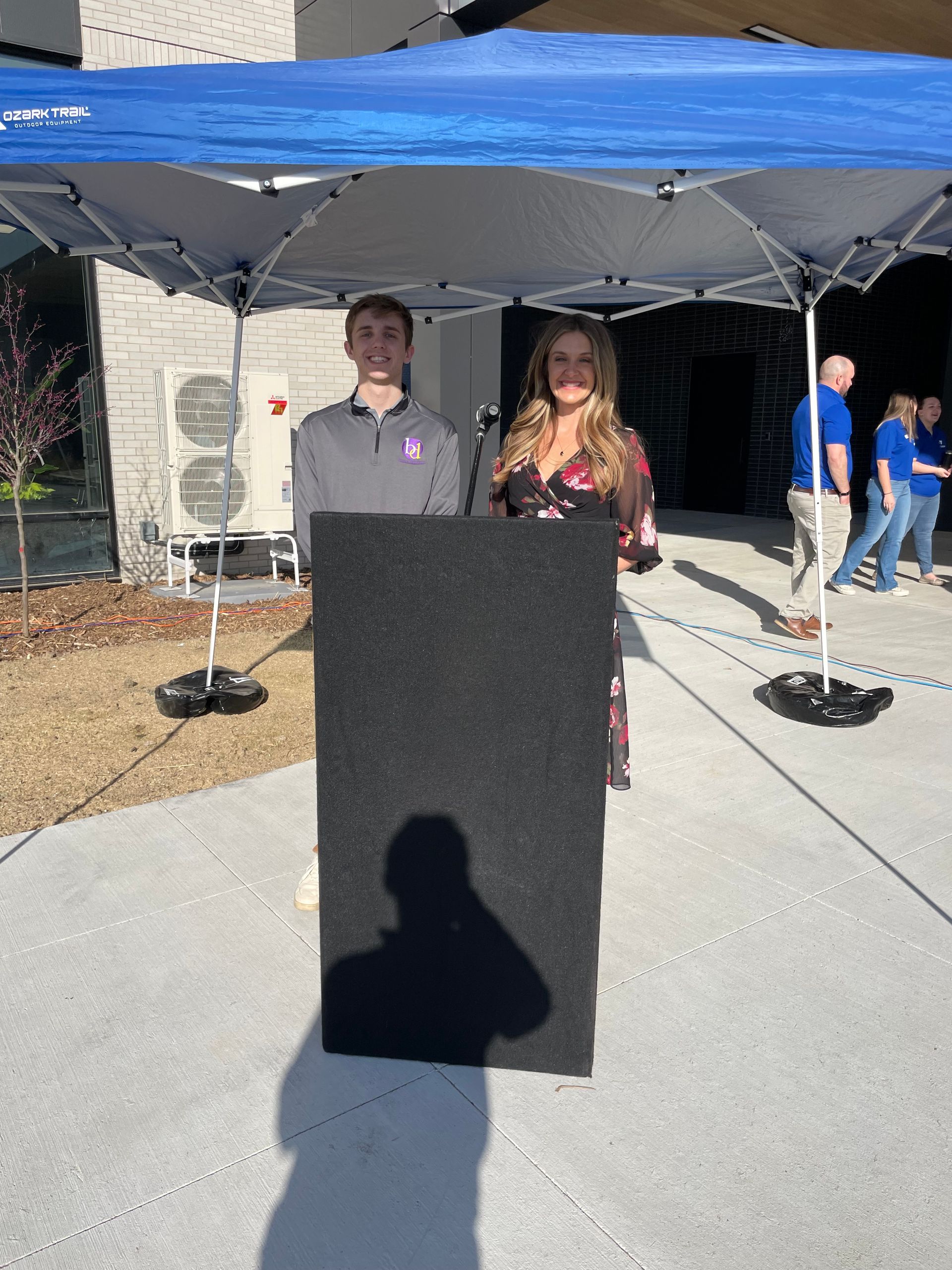 A man and a woman are standing behind a podium under a blue tent.