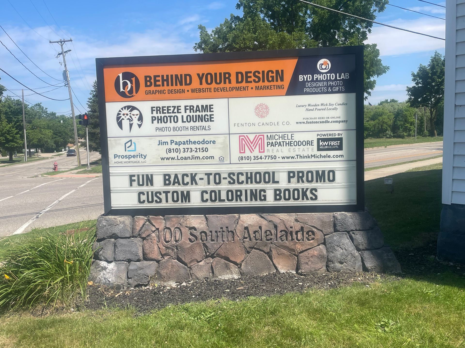 Outdoor sign for “Behind Your Design” and “Freeze Frame Photo Lounge” on a stone base in South Adelaide.