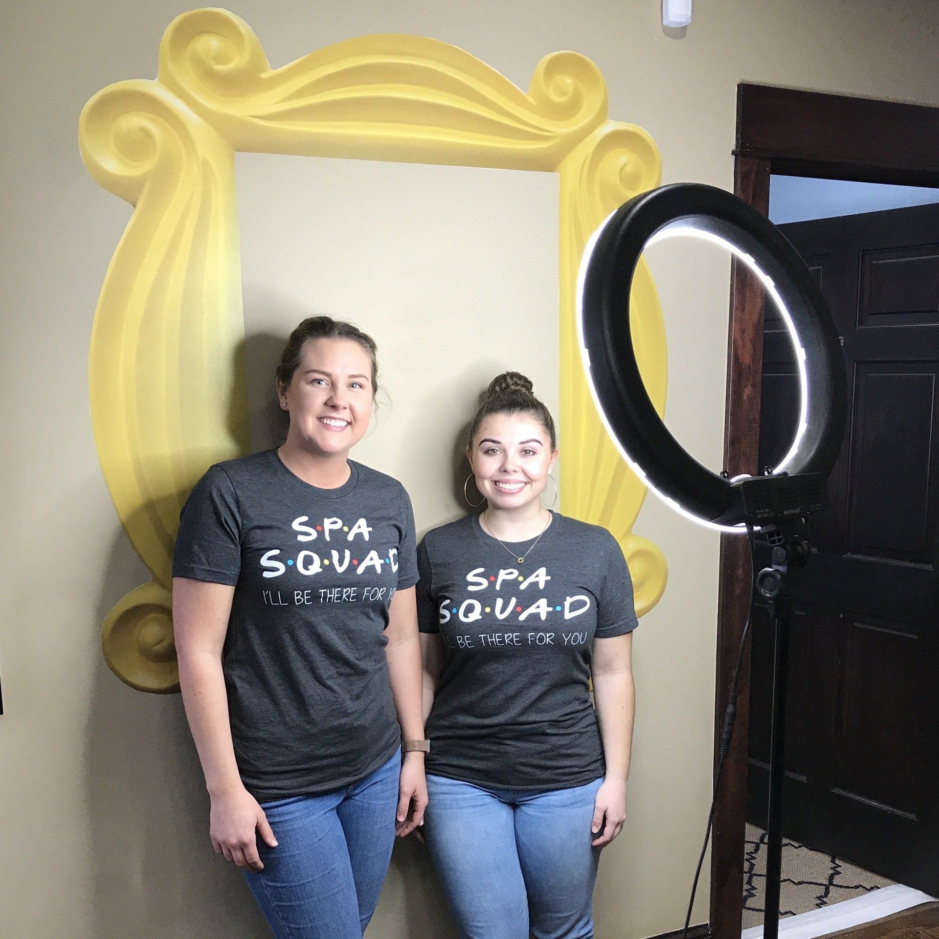 Two women wearing spa squad shirts are standing in front of a yellow frame.