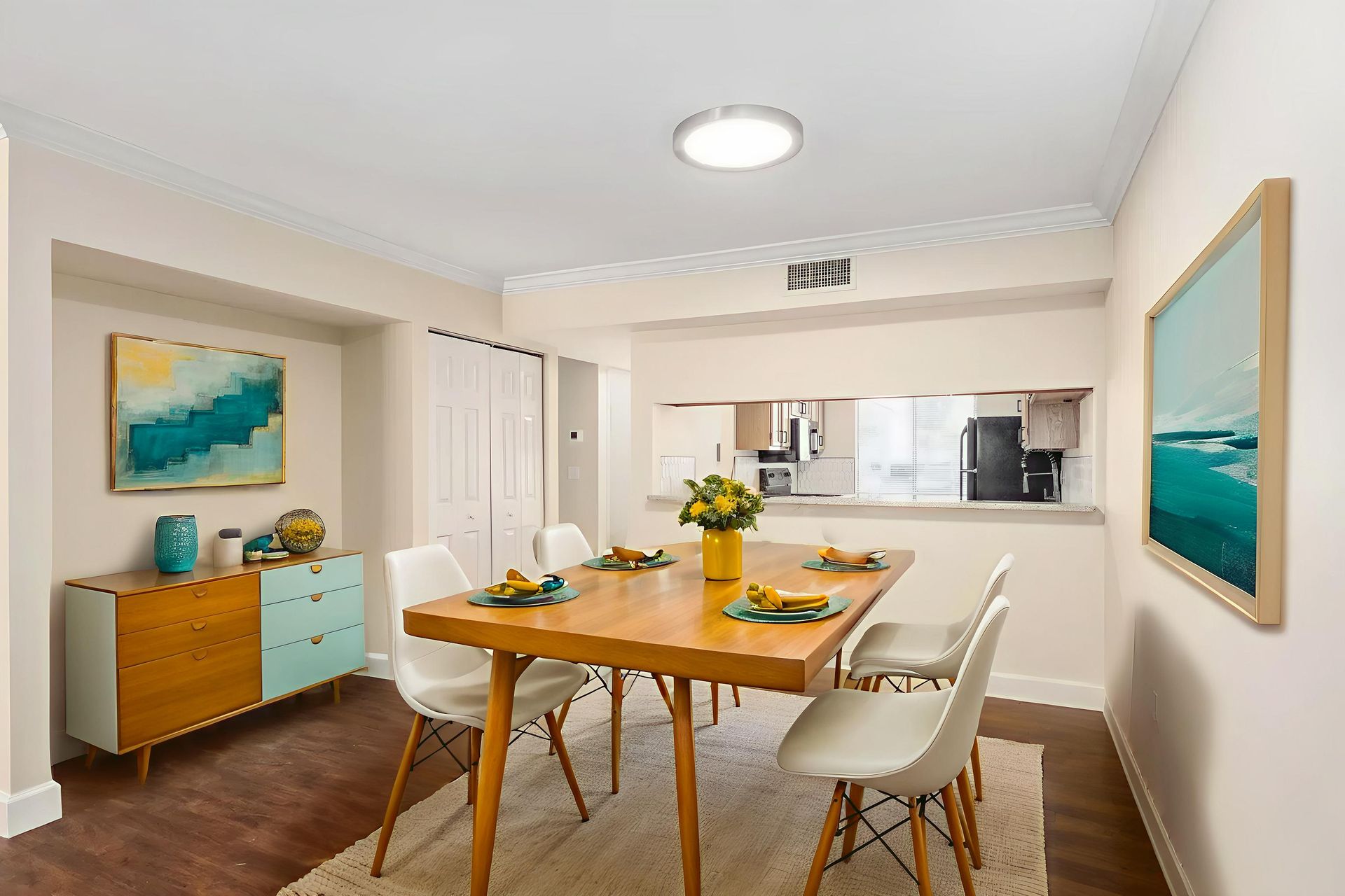Dining room with wooden table, white chairs, and colorful art. Light-colored walls with dark wood floors.