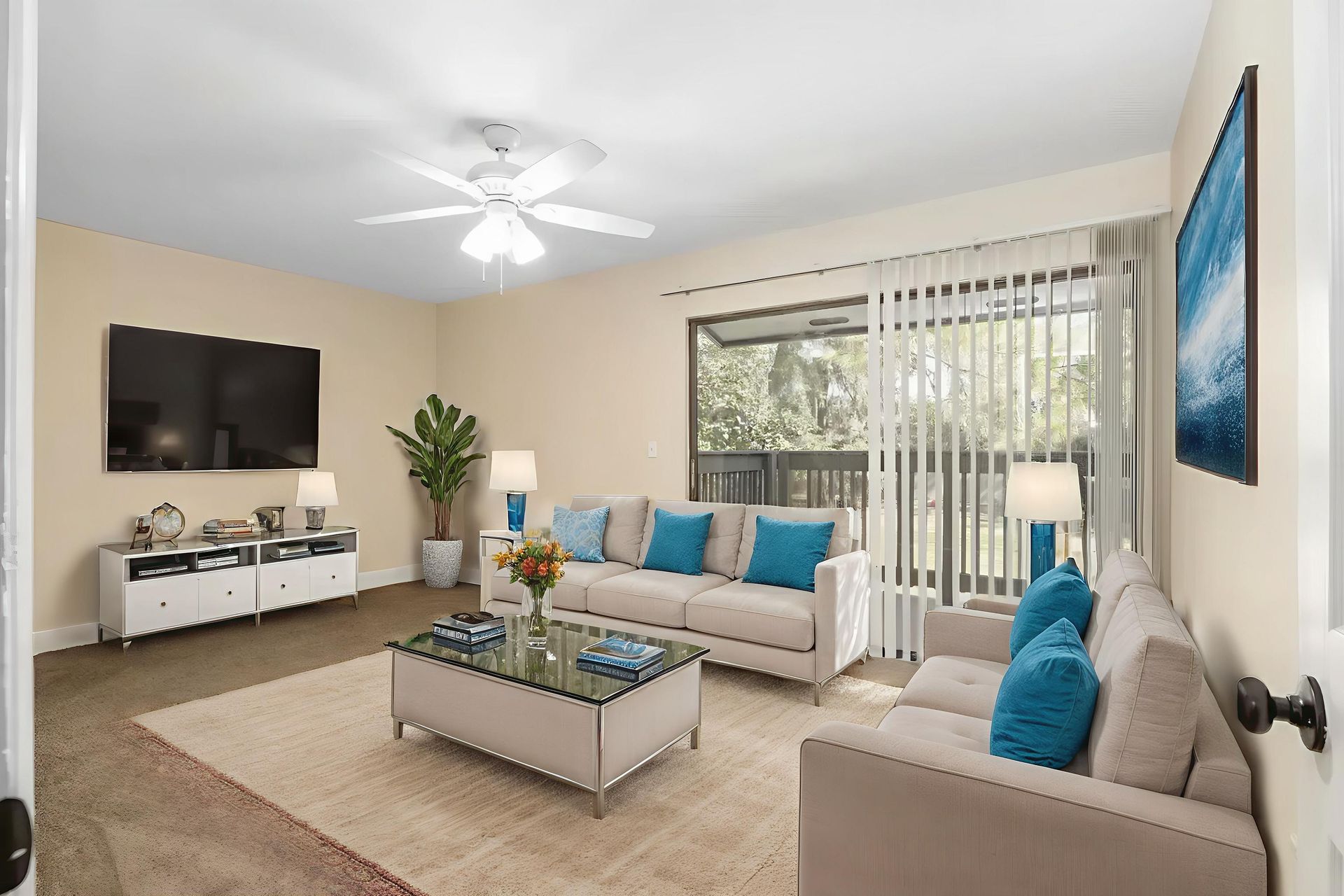Living room with beige walls, sofas with blue pillows, glass coffee table, and large TV.