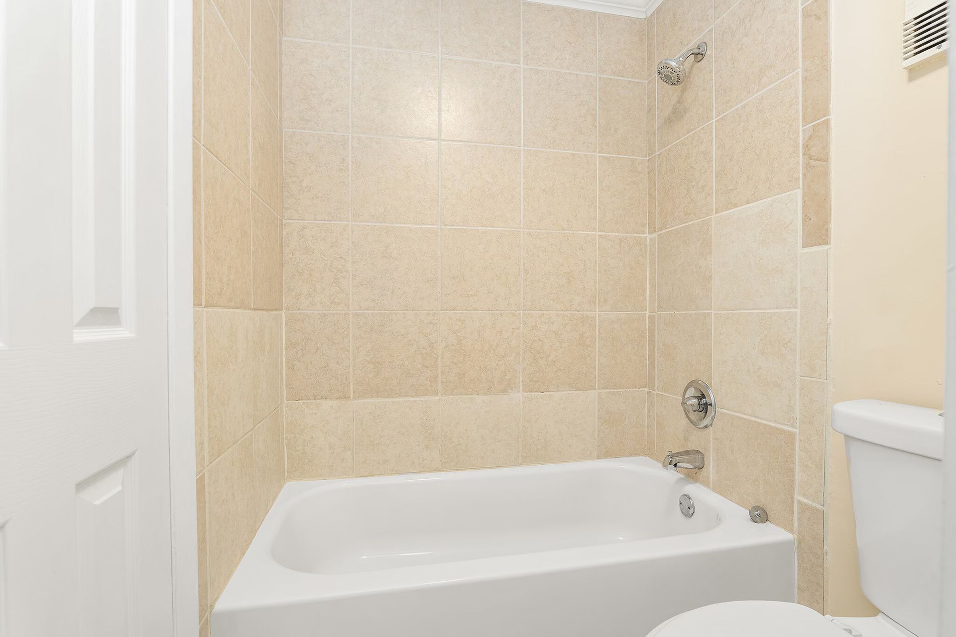 Bathroom with beige tile walls, white tub and toilet, and silver shower fixtures.