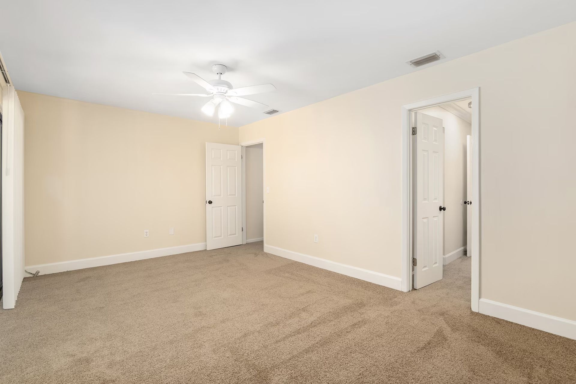 Empty bedroom with beige carpet and walls, three white doors, and a ceiling fan.
