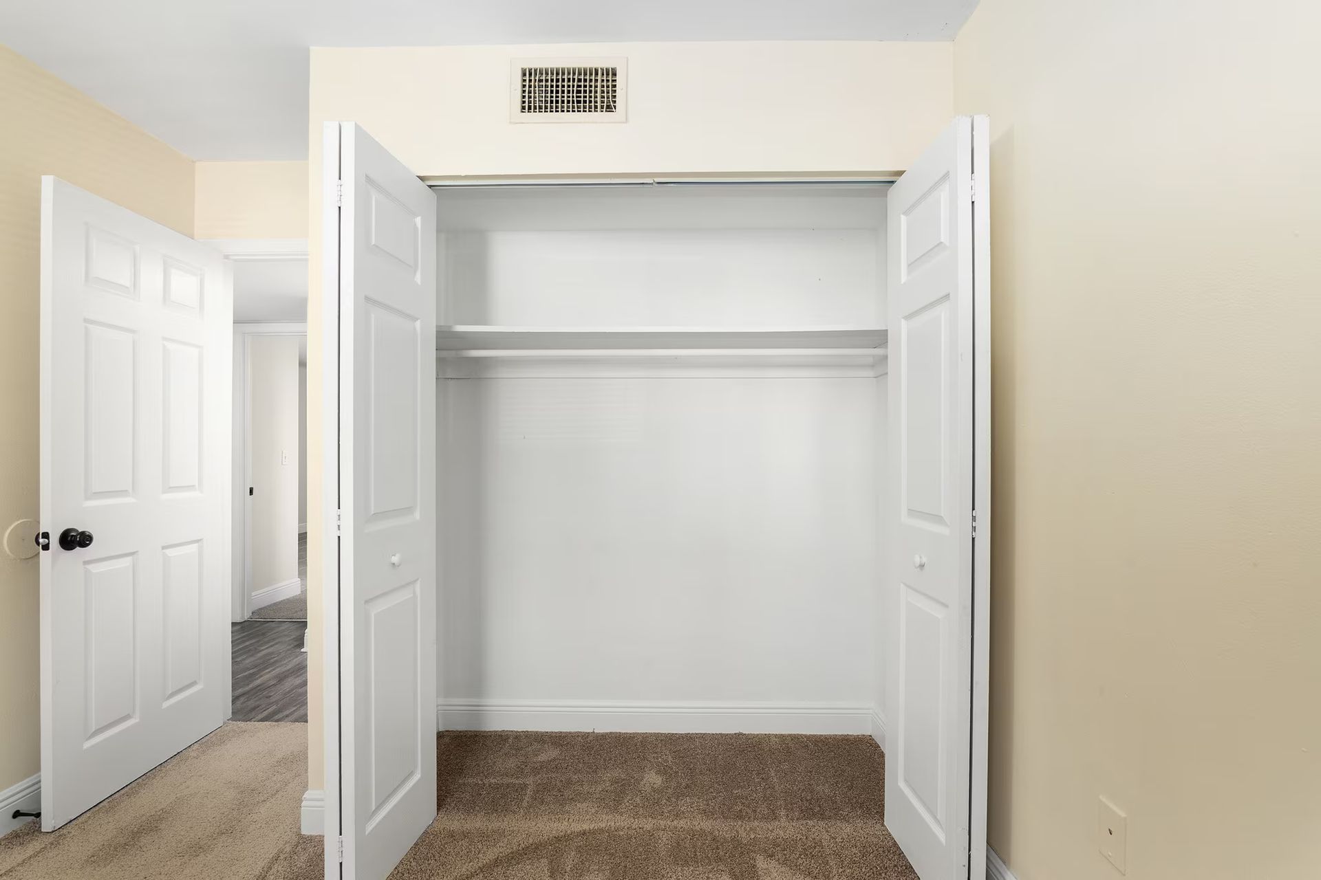 Empty closet with white bi-fold doors open, tan carpet, beige walls, and an open doorway to a hallway.
