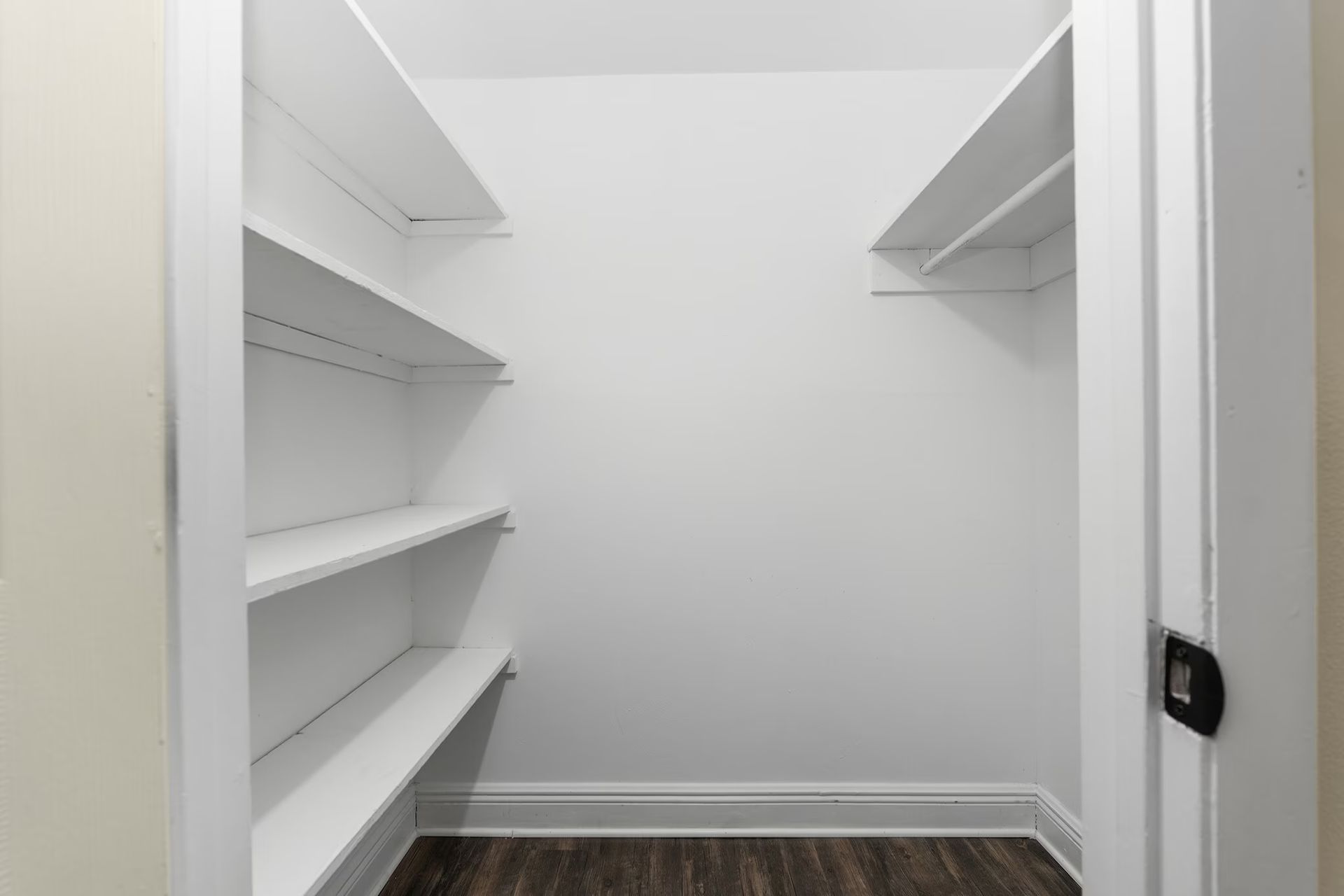 Empty white-walled closet with shelving on both sides and dark wood floor.