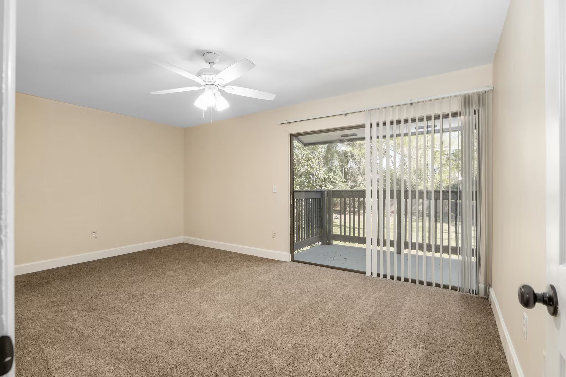 Empty bedroom with tan carpet, sliding glass door to a balcony, and a ceiling fan.