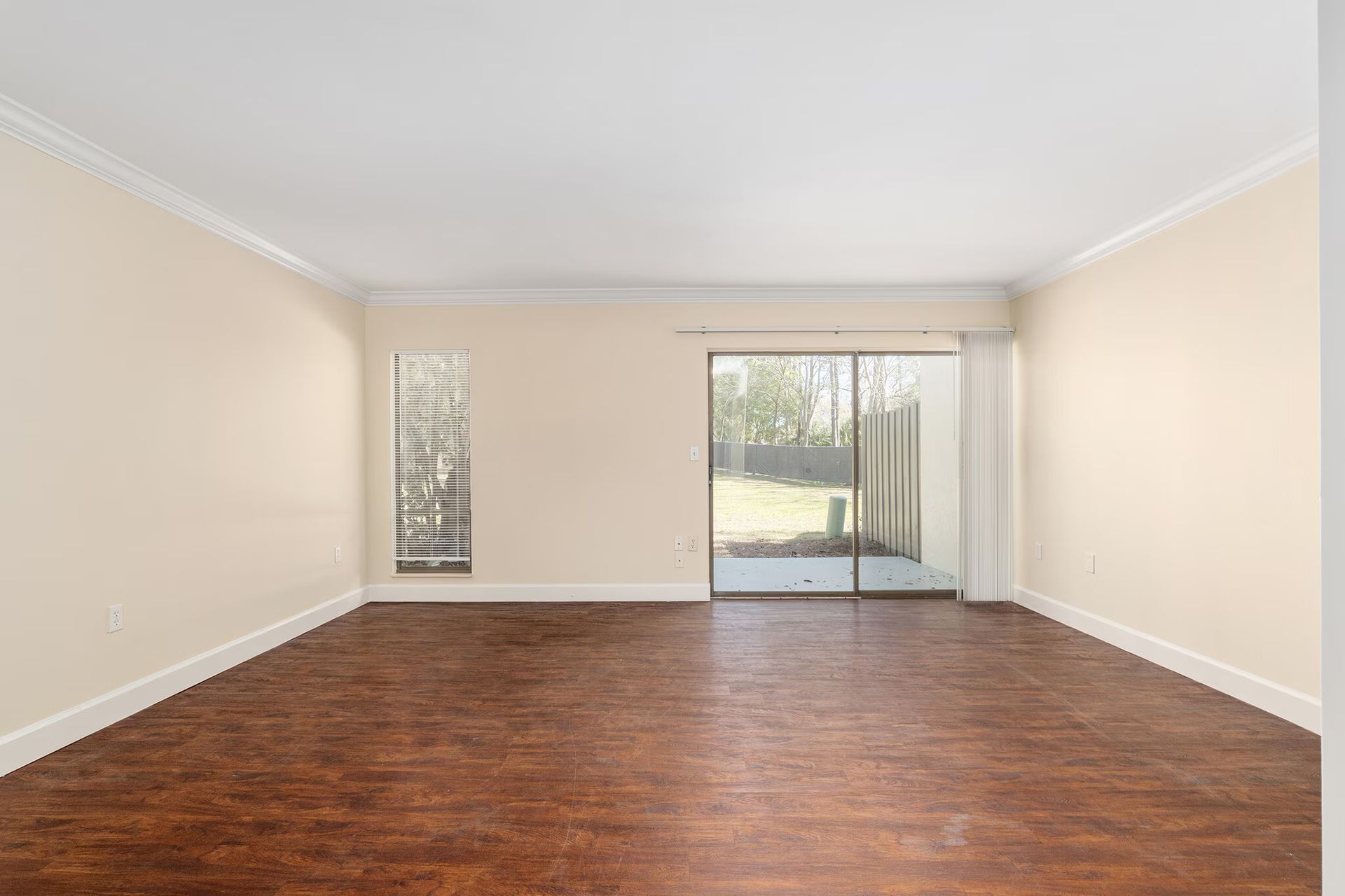 Empty living room with dark wood floor, beige walls, and sliding glass door to backyard.
