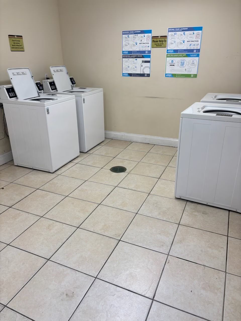 A laundry room with multiple white washing machines and tiled flooring. Instructional signs hang on the wall.
