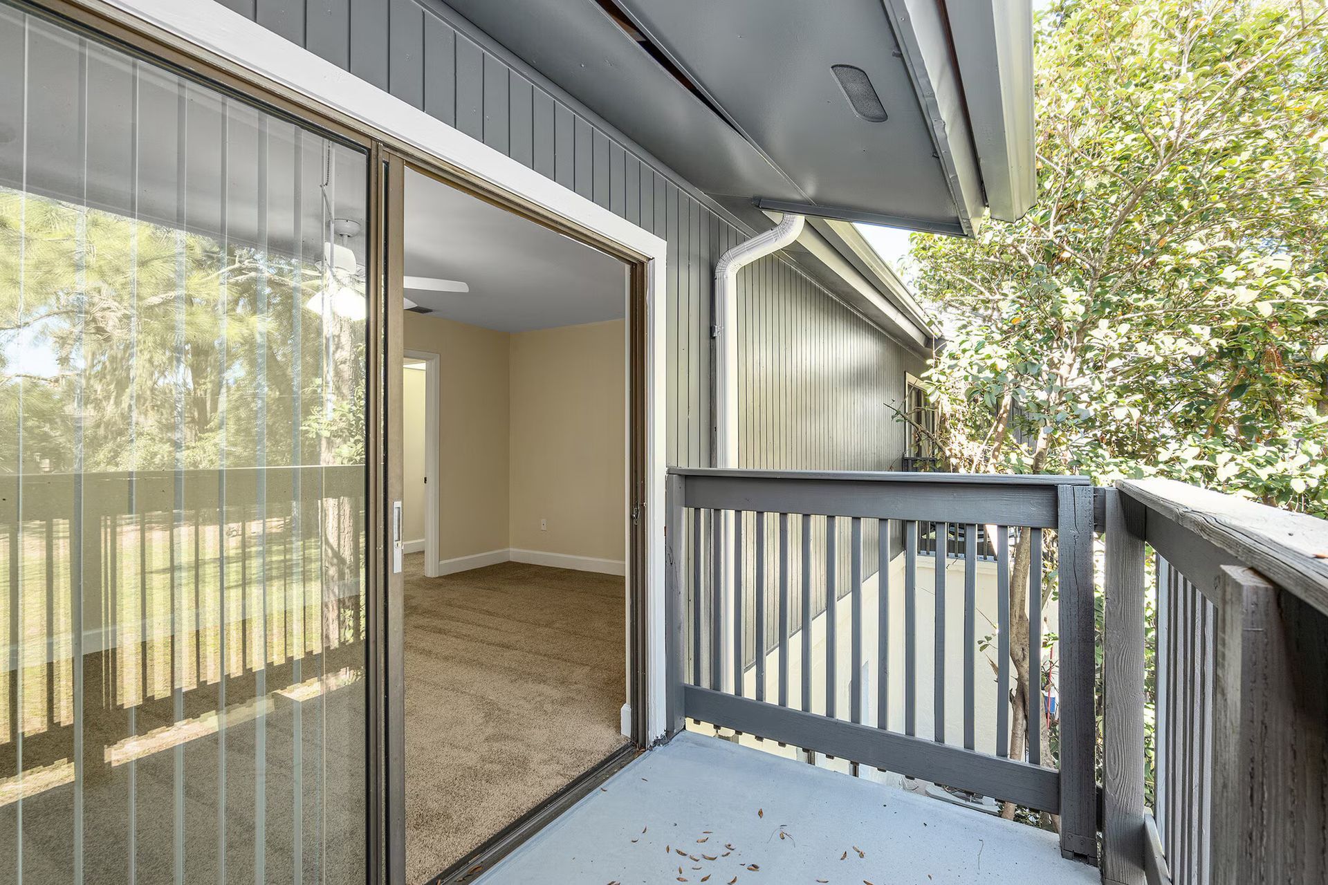 Balcony with open sliding door to a room; gray siding, brown carpet and railing, view of trees.