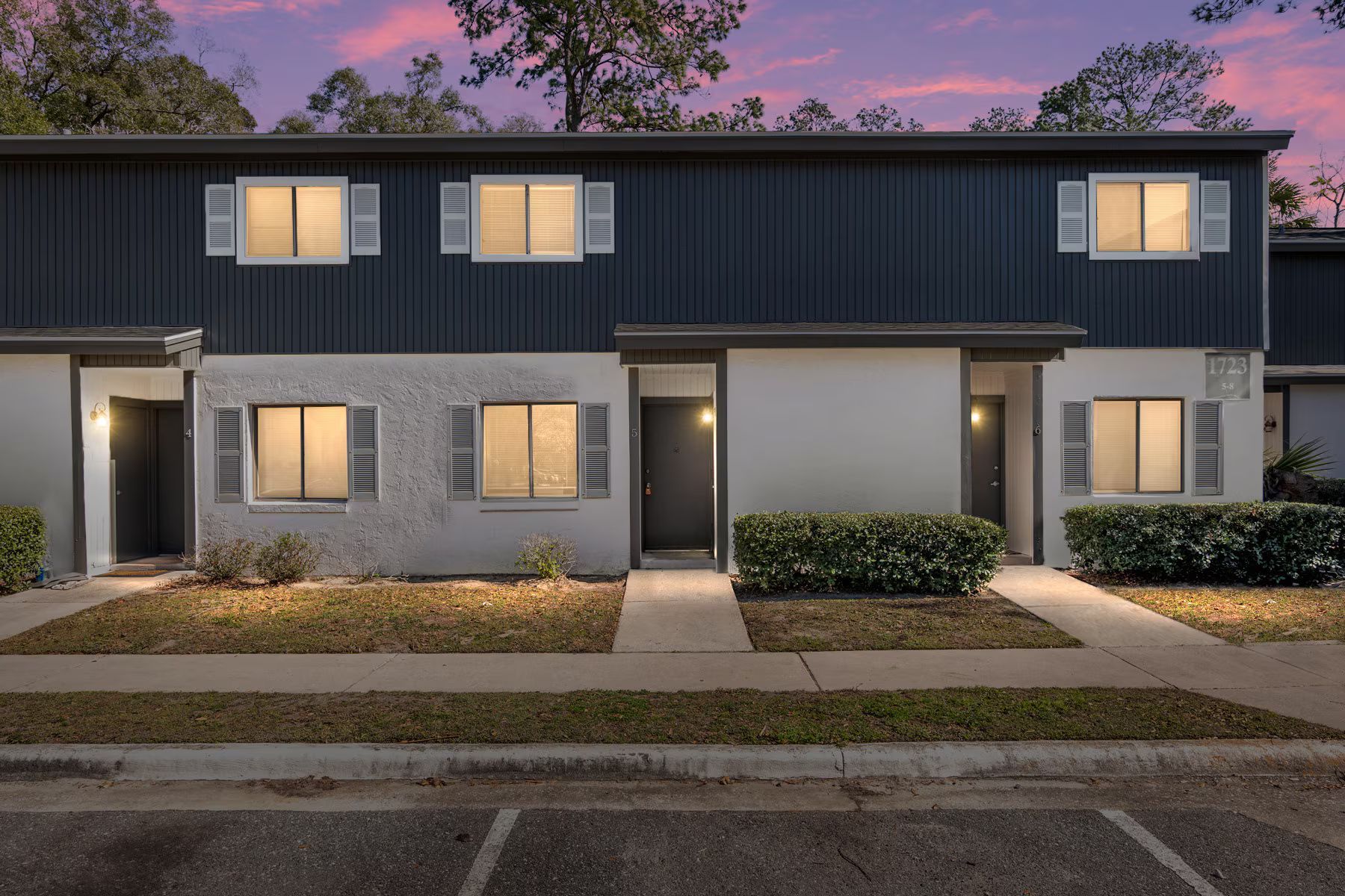 Row of townhouses with blue and gray siding, shutters, and lit windows; dusk setting.